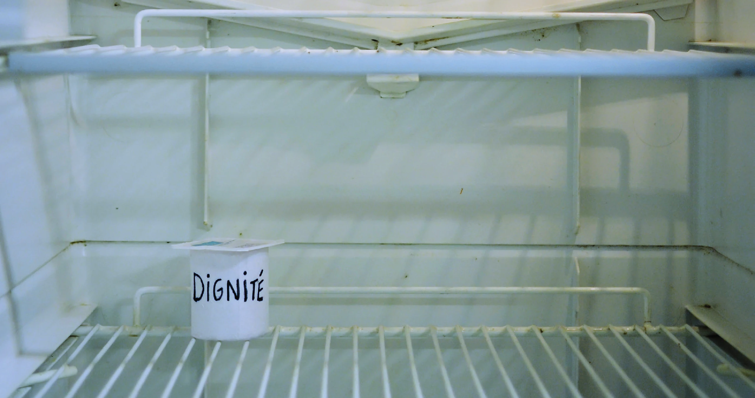 Un réfrigérateur vide avec seulement un pot de yaourt sur lequel l’étiquette est remplacée par le mot «dignité».